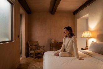 Chambre d'hôtel chaleureuse avec lampe de chevet et cliente satisfaite assise sur le lit en lin blanc